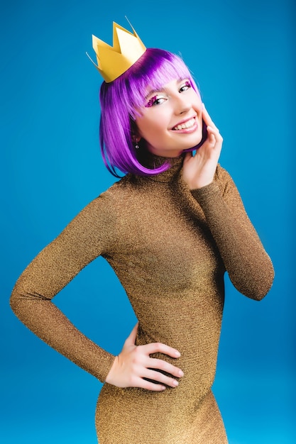 Mulher jovem elegante encantadora em vestido de ouro luxuoso sorrindo. Corte o cabelo roxo, coroa, sorriso amigável, expressando positividade, felicidade, comemoração de carnaval, festa.