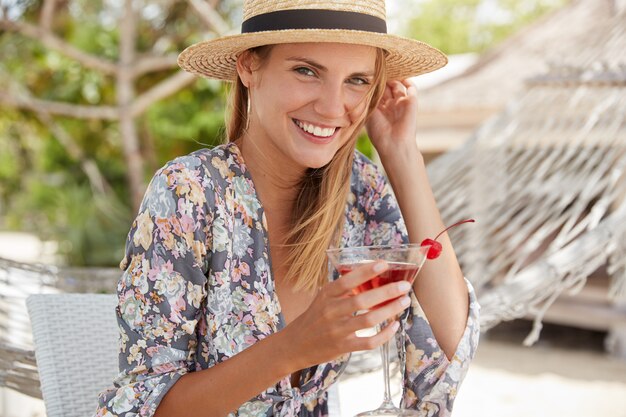 Mulher jovem elegante usa chapéu de palha de verão e camisa da moda, mantém o coquetel de cereja fresca na mão, feliz por passar o tempo livre no café ao ar livre. Mulher adorável com bebida gelada e suculenta posa do lado de fora