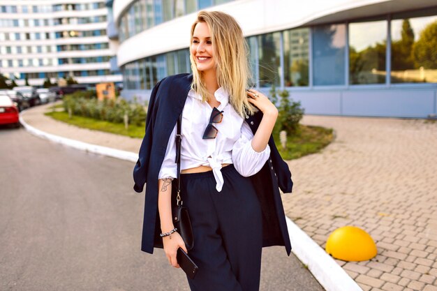 Mulher jovem elegante vestindo um terno marinho da moda, posando perto de edifícios modernos, acessórios de moda, sorrindo, aproveitando o dia ensolarado de verão, andando perto do escritório.