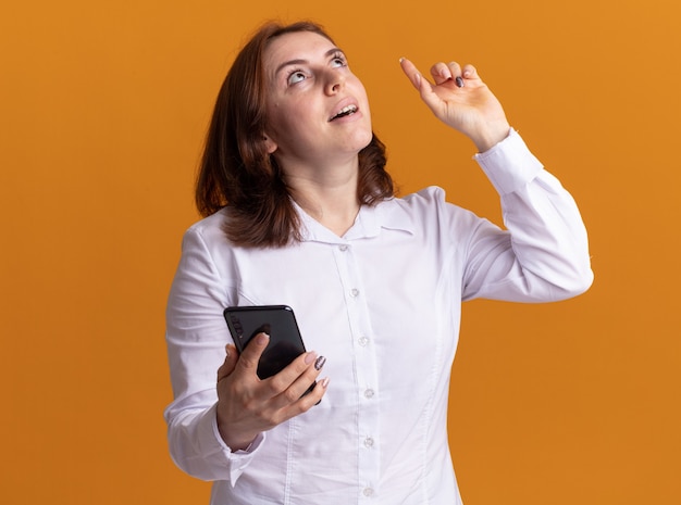 Mulher jovem em camisa branca com smartphone olhando para cima com um sorriso no rosto mostrando o dedo indicador, tendo uma nova ideia em pé sobre uma parede laranja