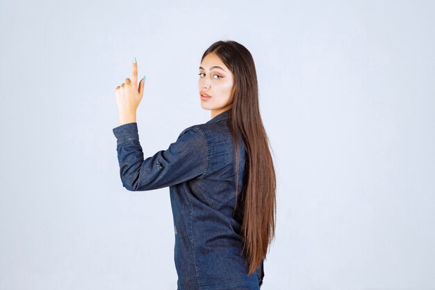 Mulher jovem em camisa jeans levantando as mãos e apontando para algo acima