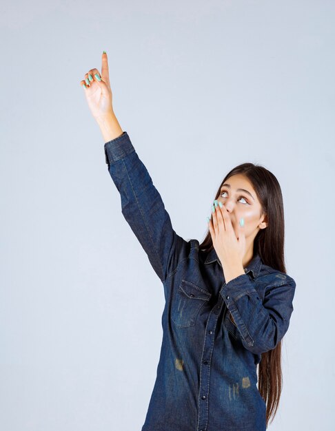 Mulher jovem em camisa jeans levantando as mãos e apontando para algo acima