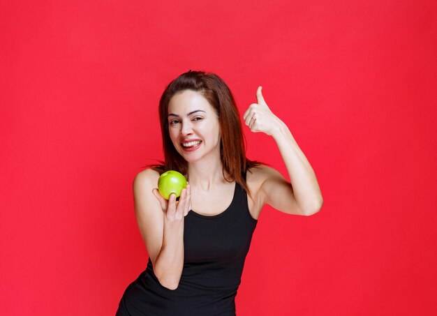 Mulher jovem em camiseta preta segurando maçãs verdes e mostrando o polegar