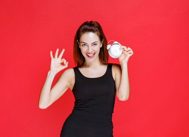 Mulher jovem em camiseta preta segurando um despertador e mostrando um sinal positivo com a mão