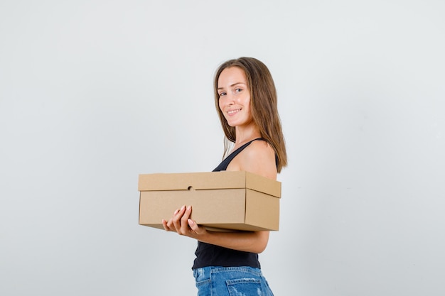 Mulher jovem em camiseta, shorts segurando uma caixa de papelão e parecendo alegre.