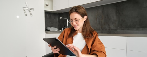 Foto grátis mulher jovem em casa lendo em óculos em tablet digital e sorrindo sentada na cozinha