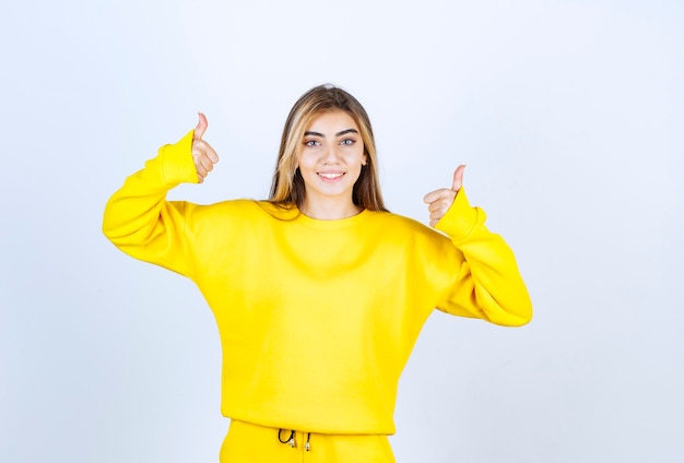 Foto grátis mulher jovem em moletom amarelo acenando com o polegar para cima na parede branca