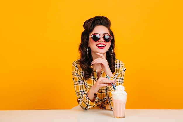 Mulher jovem em óculos de sol, bebendo milkshake. Foto de estúdio de senhora Pin-up isolada em fundo amarelo.