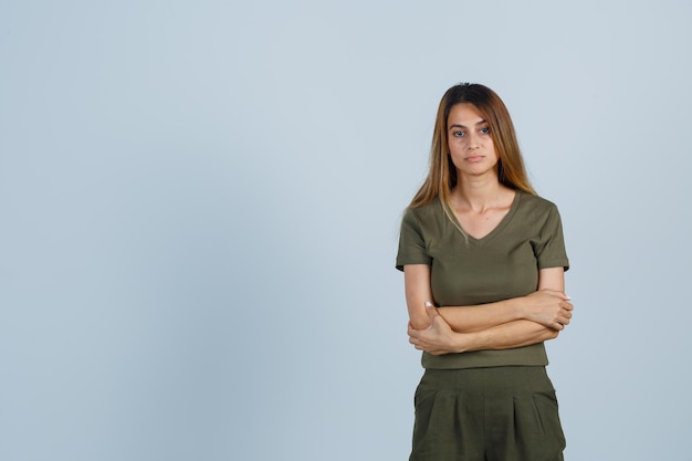 Mulher jovem em pé com os braços cruzados em t-shirt, calça e olhando triste, vista frontal.