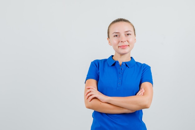 Mulher jovem em pé com os braços cruzados em uma camiseta azul e parecendo alegre