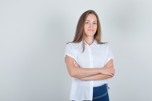 Mulher jovem em pé com os braços cruzados em uma camiseta branca, jeans e aparência alegre