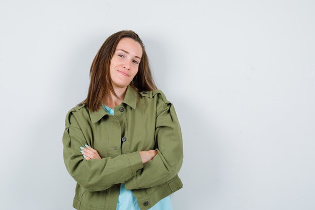 Mulher jovem em pé com os braços cruzados na jaqueta verde e olhando confiante, vista frontal.