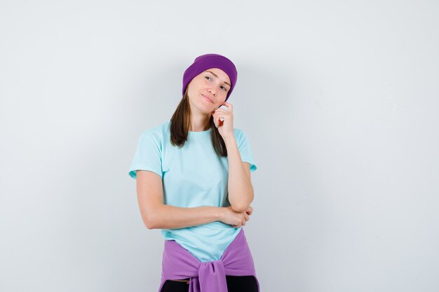 Mulher jovem em pose de pensamento, inclinando o rosto por lado na camiseta azul, gorro roxo e olhando pensativa, vista frontal.