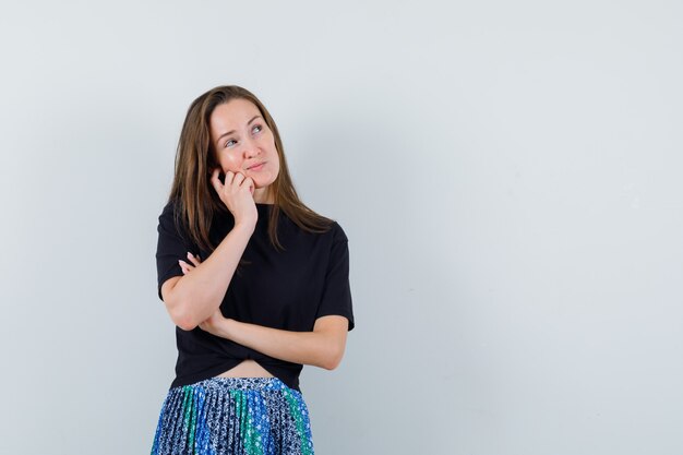 Mulher jovem em pose pensativa olhando para longe com uma camiseta preta e saia azul e parecendo pensativa