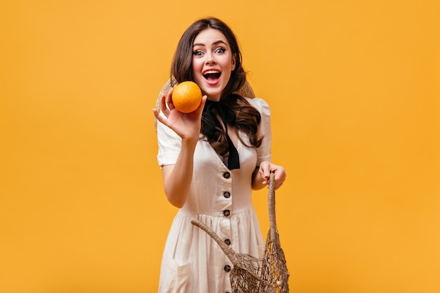 Mulher jovem em roupa branca com laço preto em volta do pescoço tira laranja de uma bolsa de barbante.