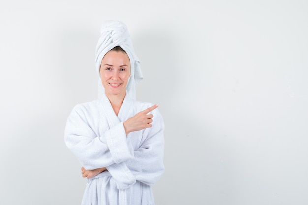 Mulher jovem em roupão branco, toalha apontando para o canto superior direito e olhando alegre, vista frontal.