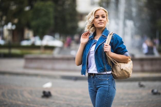 Mulher jovem em streetwalk square fontain vestida com uma suíte de jeans azul e bolsa no ombro em dia de sol