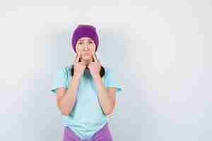 Foto grátis mulher jovem em t-shirt azul, gorro roxo dentro dex dedos perto da boca, lábios curvos e parecendo triste, vista frontal.