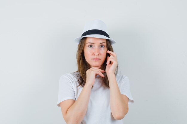 Mulher jovem em t-shirt branca, chapéu tocando a pele do rosto e parecendo tonta.