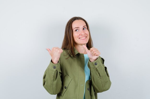 Mulher jovem em t-shirt, jaqueta apontando para o lado com os polegares e olhando alegre, vista frontal.