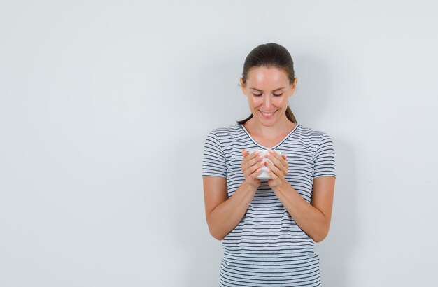 Mulher jovem em t-shirt segurando uma xícara de chá e olhando alegre, vista frontal.