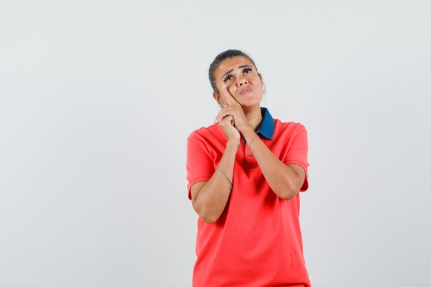 Mulher jovem em t-shirt vermelha, inclinando a bochecha por lado e olhando pensativa, vista frontal.