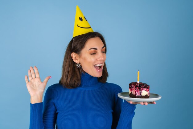 Mulher jovem em uma parede azul comemora aniversário, segura um pedaço de bolo, de ótimo humor, feliz, animada