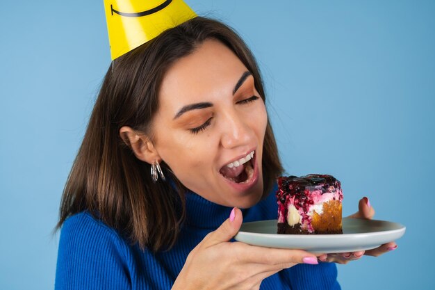 Mulher jovem em uma parede azul comemora aniversário, segura um pedaço de bolo, feliz, morde o bolo