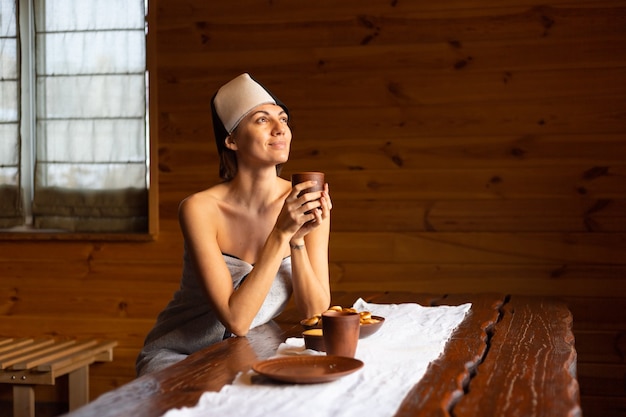 Mulher jovem em uma sauna com um boné na cabeça se senta a uma mesa e bebe chá de ervas, desfrutando de um dia de bem-estar