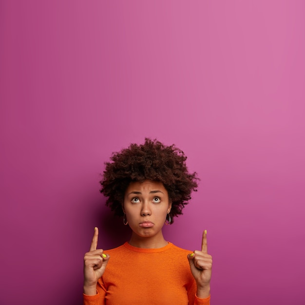 Foto grátis mulher jovem encaracolada decepcionada e chateada aponta para cima com os dedos indicadores, olha com tristeza e desânimo acima, tem expressão abatida, vestida com roupas casuais, posa sobre uma parede amarela vibrante.