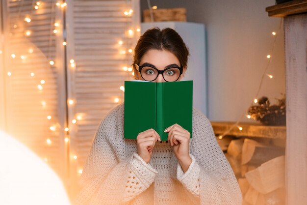 Mulher jovem engraçada com óculos na de guirlandas de luz segurando um livro nas mãos e lendo