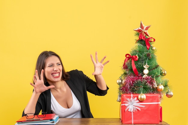 Mulher jovem engraçada olhando para algo com expressão facial de surpresa, sentada à mesa perto da árvore de Natal decorada no escritório em amarelo