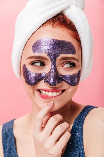 Mulher jovem espetacular fazendo tratamento de spa com um sorriso sincero. Foto de estúdio de garota feliz com máscara facial e toalha.