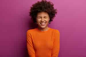 Foto grátis mulher jovem étnica muito feliz ri de algo positivo, fecha os olhos e ri alegremente, vestida com um macacão laranja, ouve uma piada engraçada, posa contra uma parede roxa vibrante. conceito de emoções