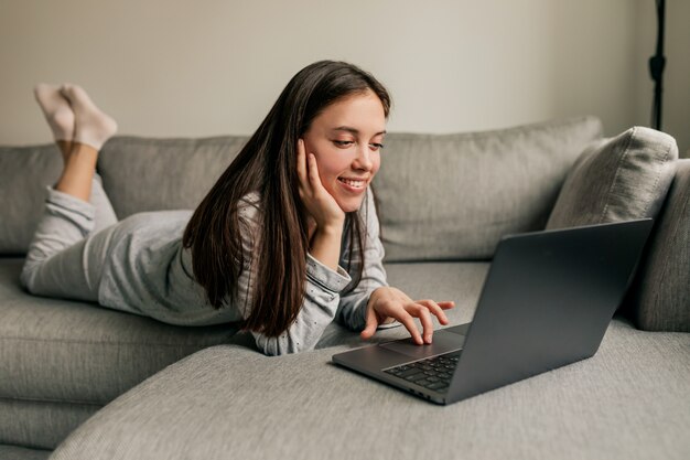 Mulher jovem europeia atraente com longos cabelos escuros de pijama, trabalhando em casa com o laptop durante a quarentena.