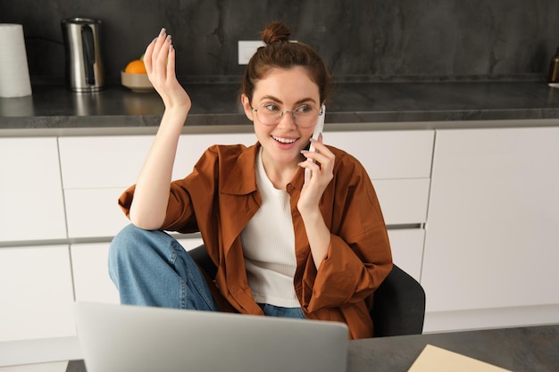 Foto grátis mulher jovem excitada falando em celular na frente do laptop sentada na cozinha com rosto feliz