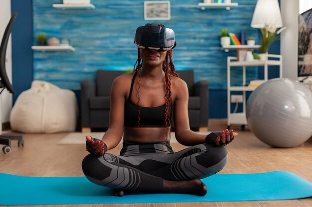 Mulher jovem experimentando realidade virtual, treinando corpo e mente, meditando na posição de lótus, sentada no tapete de ioga na sala de estar de sua casa