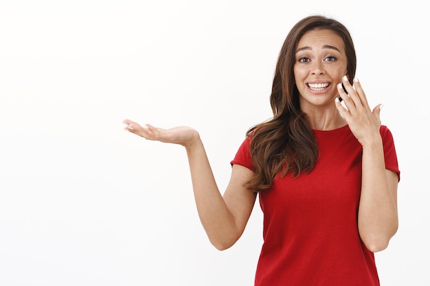 Mulher jovem extremamente feliz e lisonjeada recebe prêmio, parece sentimental e agradecida, sorrindo animada, segurando algo na palma da mão e enxugando uma lágrima de alegria do rosto, parede branca