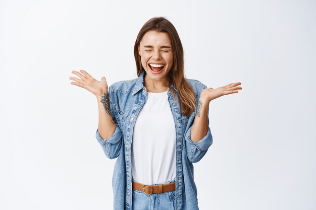 Mulher jovem extremamente feliz gritando de felicidade e alegria, vencendo e comemorando, gritando de alegria, em pé contra uma parede branca alegre