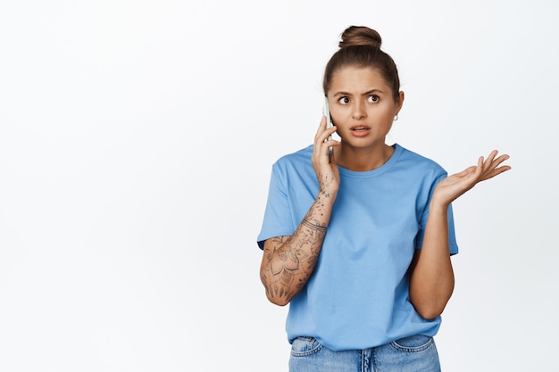 Foto grátis mulher jovem falando ao celular com cara confusa, em pé com uma camiseta azul sobre branco