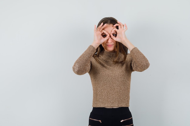 Foto grátis mulher jovem fazendo gesto binocular no olho com uma blusa dourada e parecendo esquisito