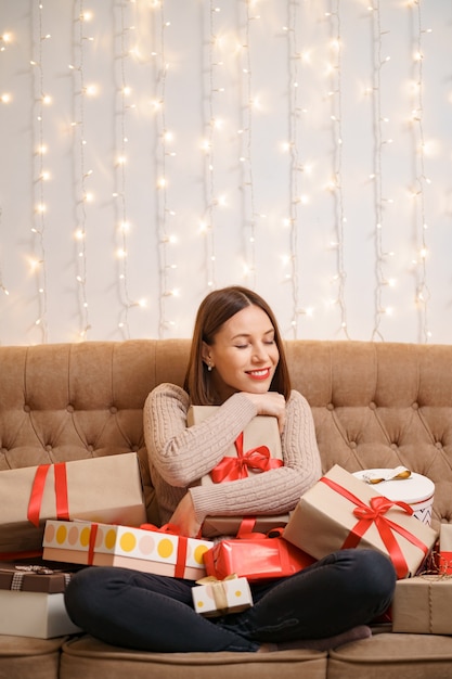 Mulher jovem feliz abraçando muitas caixas de presentes, sentada de pernas cruzadas em um sofá de camelo com luzes