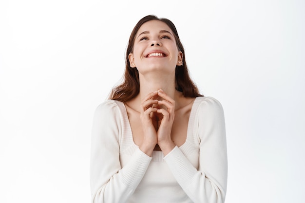 Mulher jovem feliz agradecendo a deus, parecendo aliviada e alegre por cima, dizendo orações, expressando gratidão e alegria, fazendo desejo, regozijando-se em pé contra a parede branca