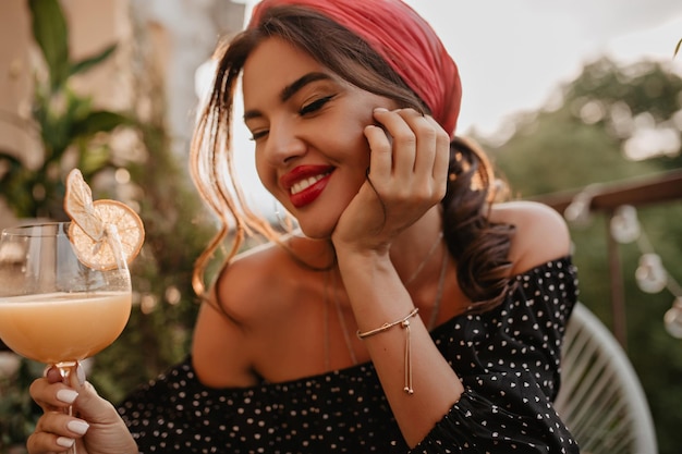 Mulher jovem feliz com bandana rosa de cabelo moreno e sorriso bonito na camisa de bolinhas de manga comprida sorrindo e posando com copo de coquetel