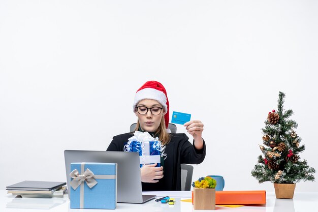 Mulher jovem feliz com chapéu de papai noel e usando óculos, sentada à mesa segurando um presente de natal e cartão do banco.