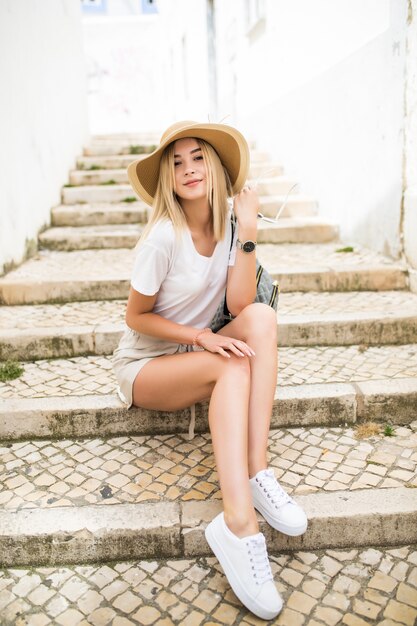 Mulher jovem feliz com chapéu de verão sentada na escada na rua da cidade