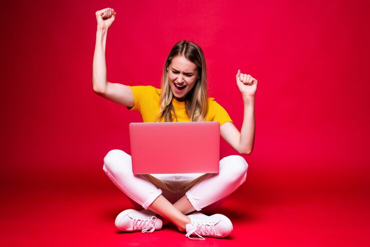 Foto grátis mulher jovem feliz com gesto vencedor, sentado no chão com as pernas cruzadas e usando o laptop na parede vermelha.