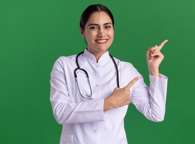 Mulher jovem feliz com jaleco com estetoscópio olhando para frente com um sorriso no rosto apontando com o dedo indicador para o lado em pé sobre a parede verde