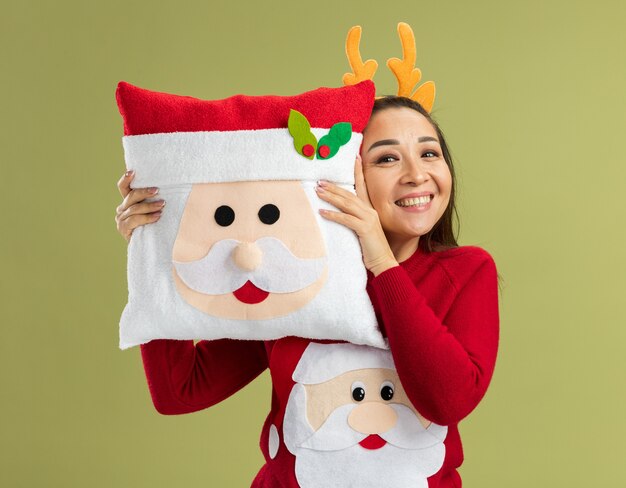 Mulher jovem feliz com um suéter vermelho de Natal, vestindo uma borda engraçada com chifres de veado segurando uma almofada de Natal e sorrindo alegremente em pé sobre a parede verde