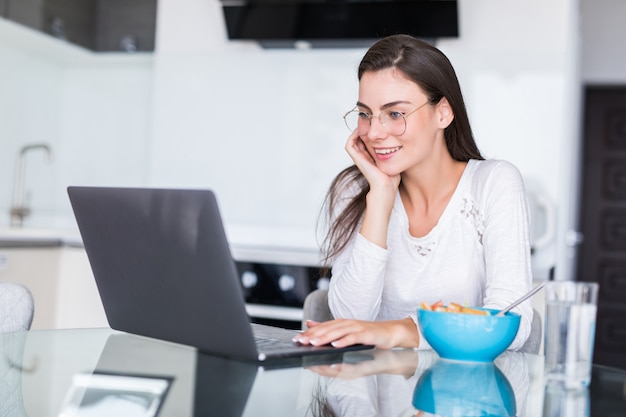 Mulher jovem feliz comendo salada de uma tigela e bebendo suco de laranja em pé em uma cozinha e assistindo filme no laptop